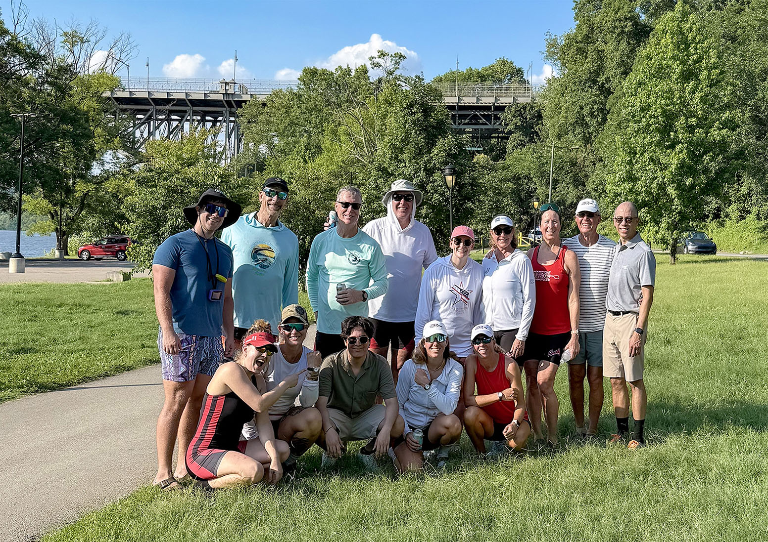 JRC IDR 2025 2 - Edit Web JRC racers posing in a group shot, standing on grass next to a river, with trees and a bridge in the background